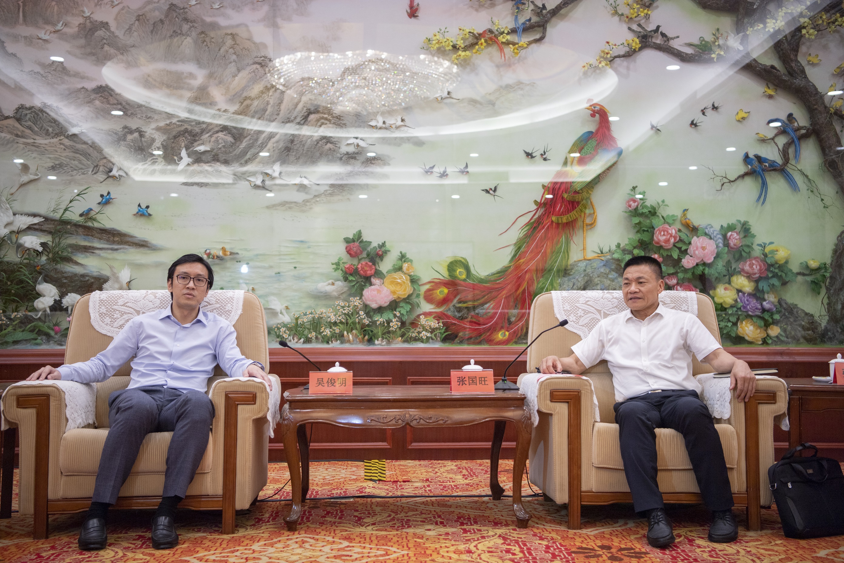 Two men in armchairs face forward, tables between them, ornate wall art with birds behind.